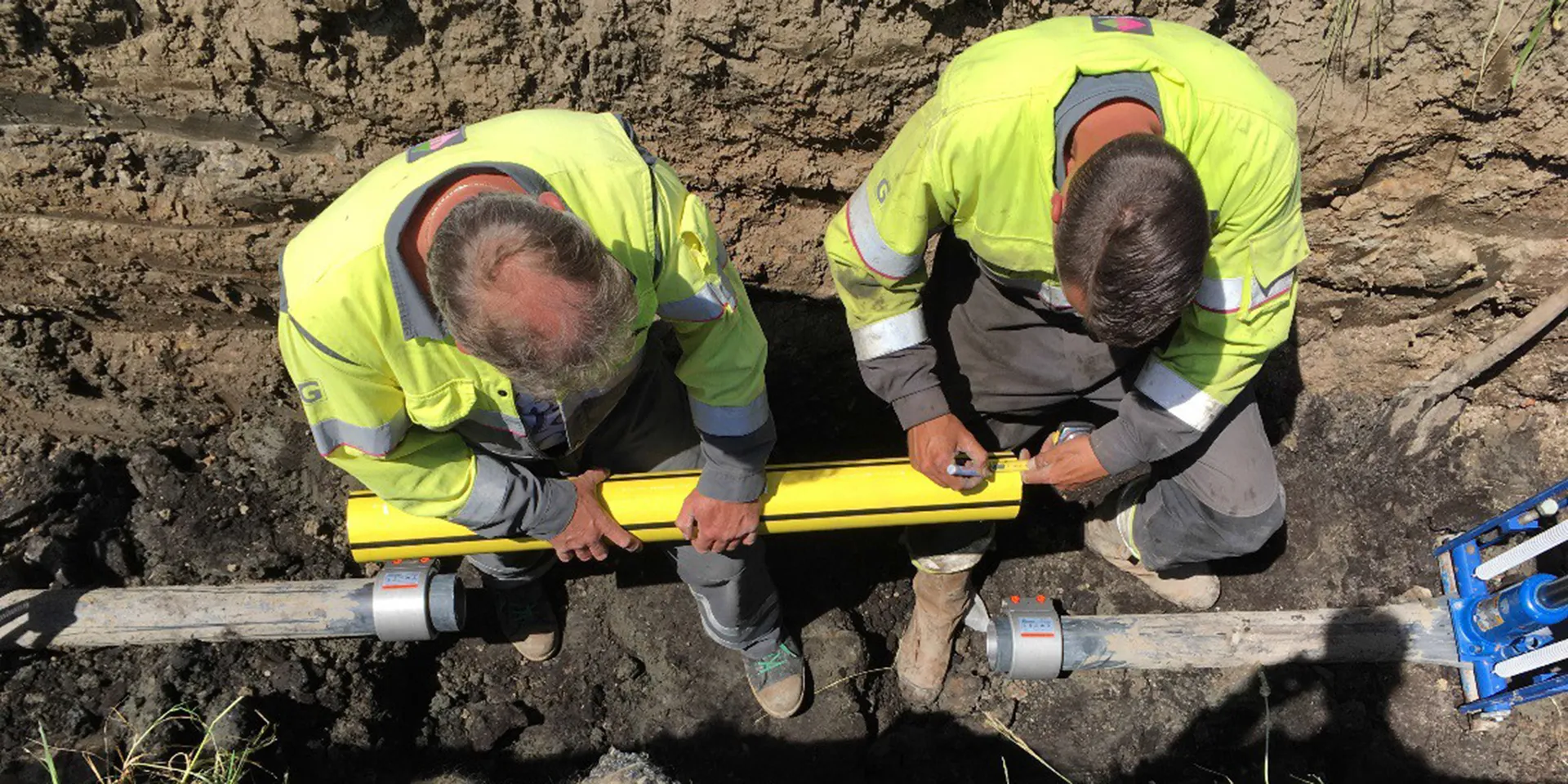 Two technicians fitting a pipe coupling to a pipeline in an excavation